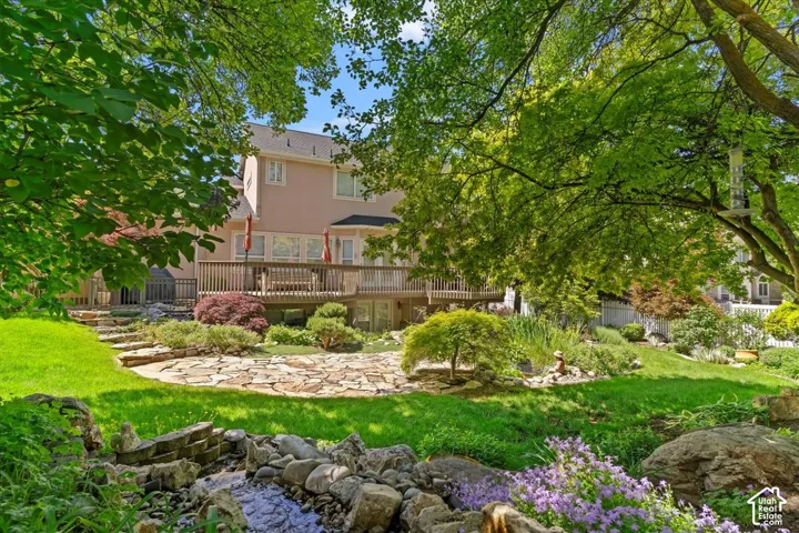 Lush green backyard featuring a multi-level wood deck, stone-paved patio, and a decorative rock-lined water feature