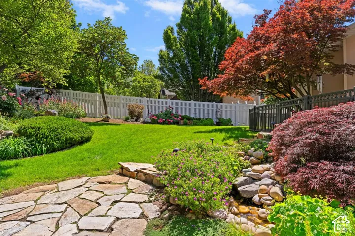 Landscaped backyard featuring a stone pathway, a manicured lawn, a white picket fence, a tiered rock garden with a water feature, and mature trees including a vibrant Japanese maple