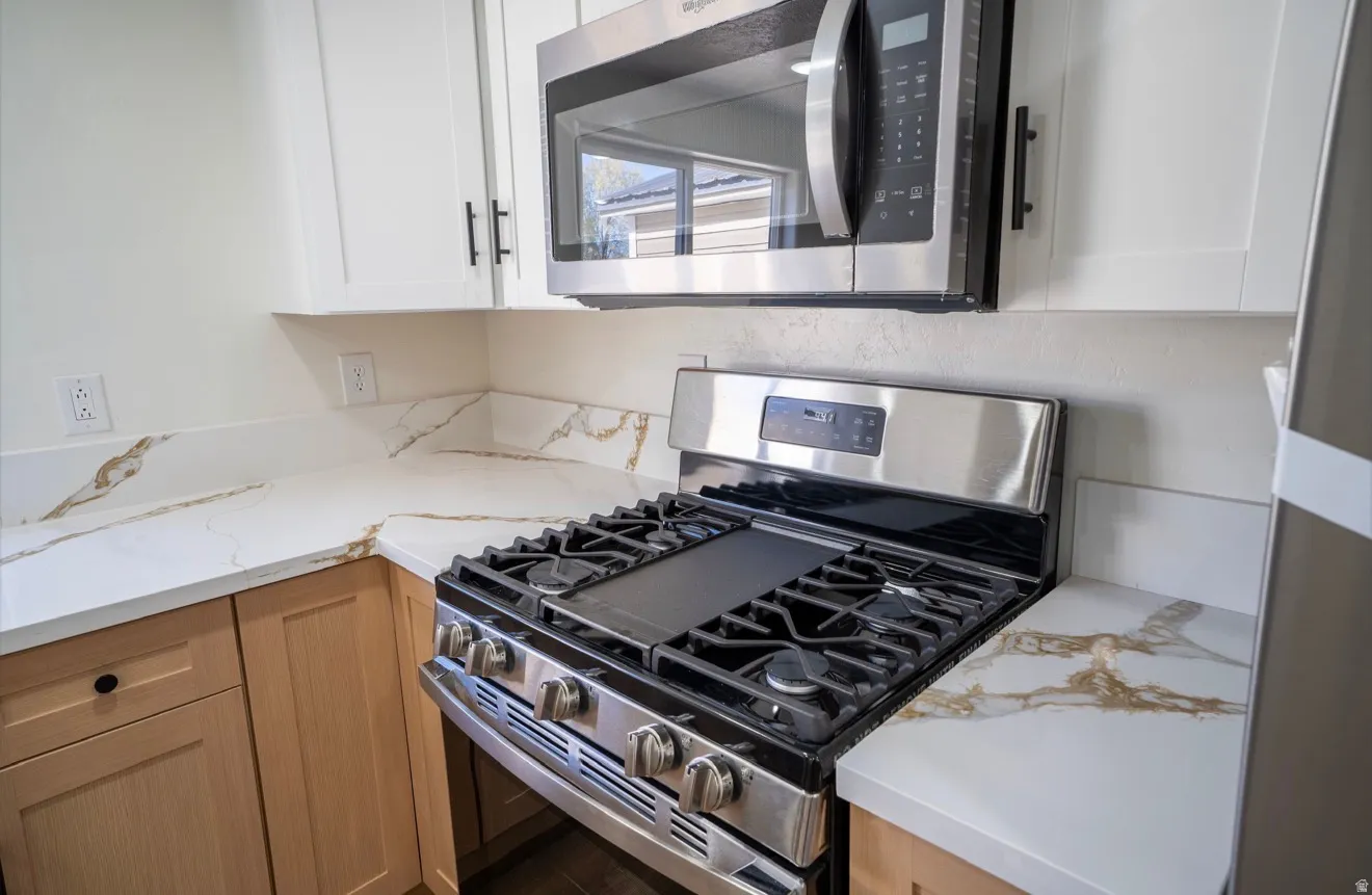 Kitchen featuring a stainless steel gas range, coordinating stainless steel microwave, white upper cabinetry with black hardware, and light wood-finish lower cabinetry, all complemented by white countertops with natural veining