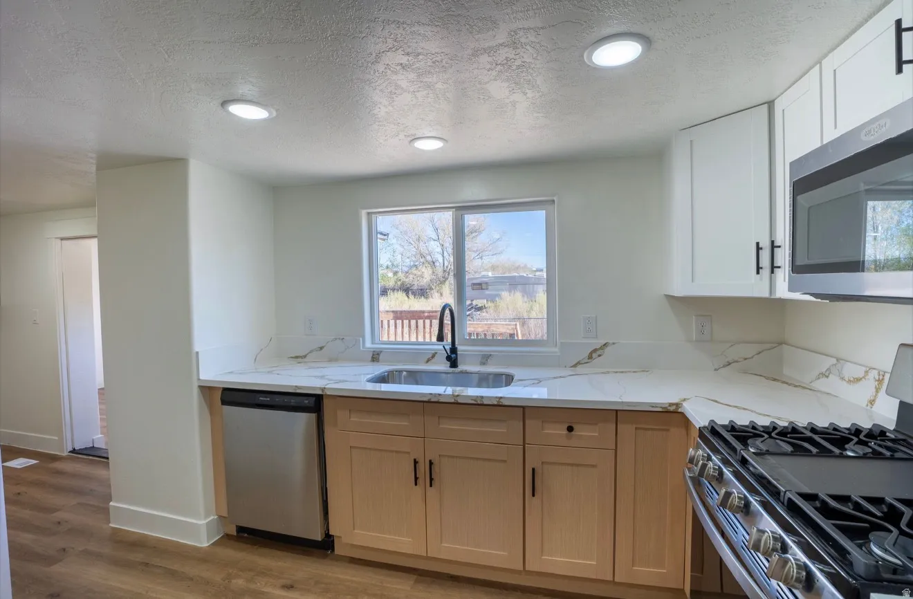 Kitchen featuring recessed lighting, a stainless steel sink with a black gooseneck faucet, light wood-finish lower cabinetry, white upper cabinetry, and a stainless steel dishwasher
