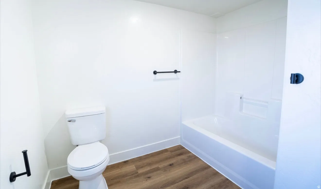 Bathroom featuring a white toilet, integrated bathtub/shower unit, wood-finish flooring, and matte black hardware