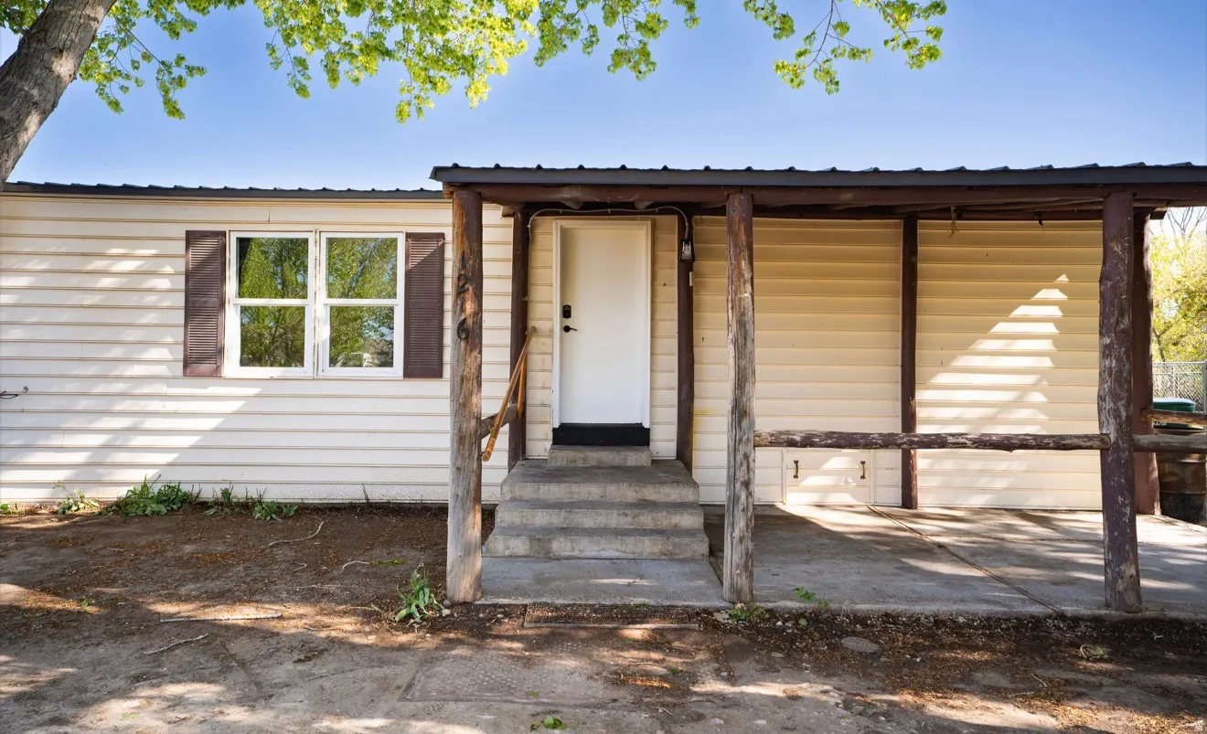 Front exterior featuring light-colored siding, a covered porch with rustic wood posts, a white entry door, concrete steps, and a window with dark shutters