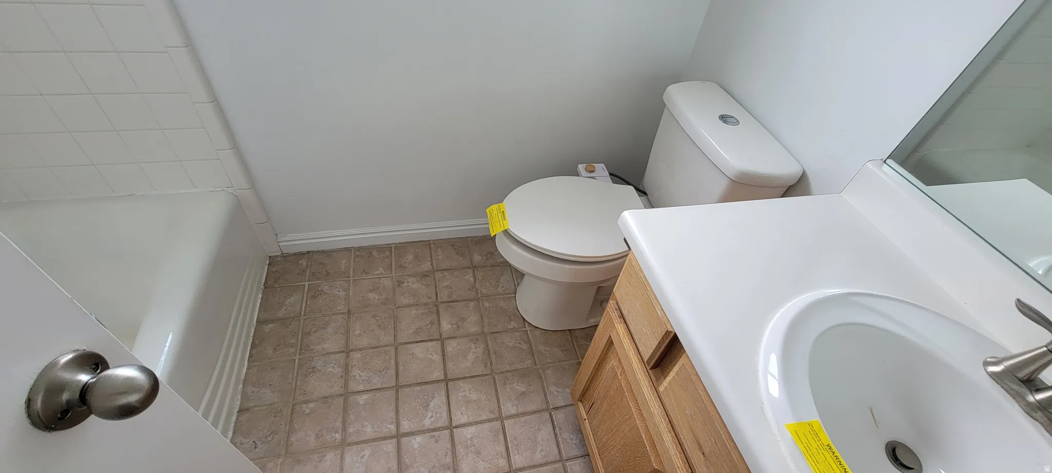 Bathroom featuring a white bathtub, light-colored square floor tiles, a standard toilet, a wood-finish vanity with a white countertop, and a built-in sink with a brushed nickel faucet