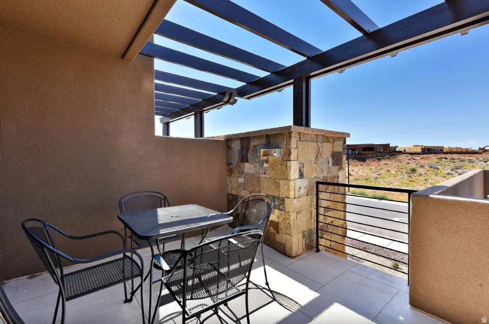 Outdoor patio featuring an open pergola, stucco walls, stone accent pillar, black metal railing, and concrete flooring