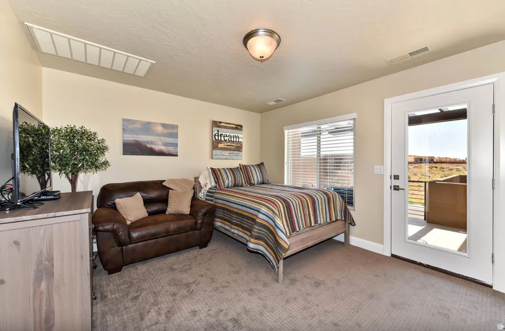 Carpeted room featuring a ceiling-mounted light fixture, a window with horizontal blinds, and a glass-paneled door opening to an outdoor patio with metal railings and a desert landscape beyond