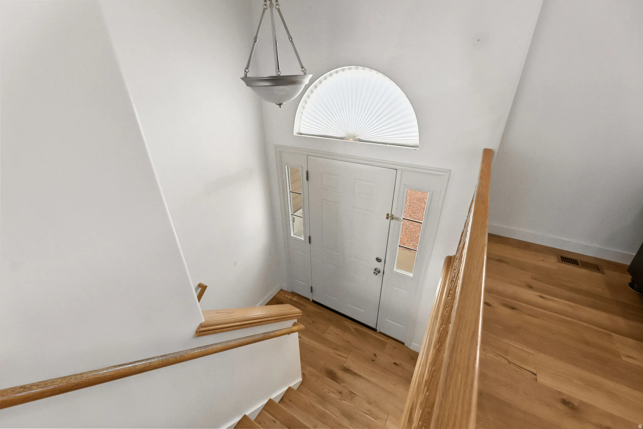 Entryway featuring wood-finish flooring, a white paneled entry door with sidelights, an arched transom window with blinds, a hanging pendant light fixture, and a light wood-finish staircase with matching handrails