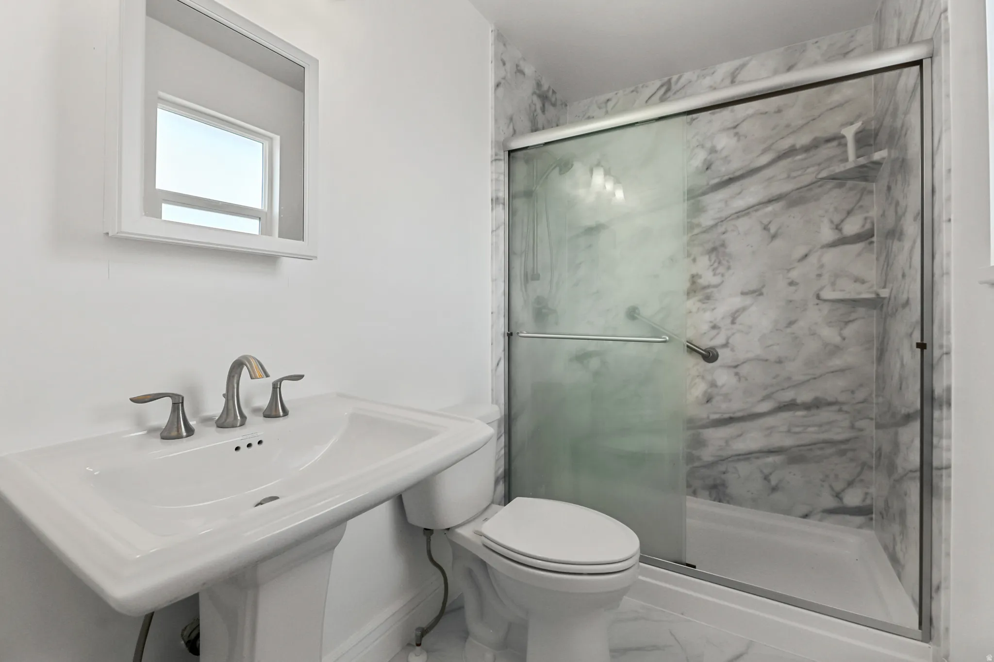 Bathroom featuring a pedestal sink with brushed nickel faucet, a toilet, and a glass-enclosed shower with marbled wall panels and built-in shelving