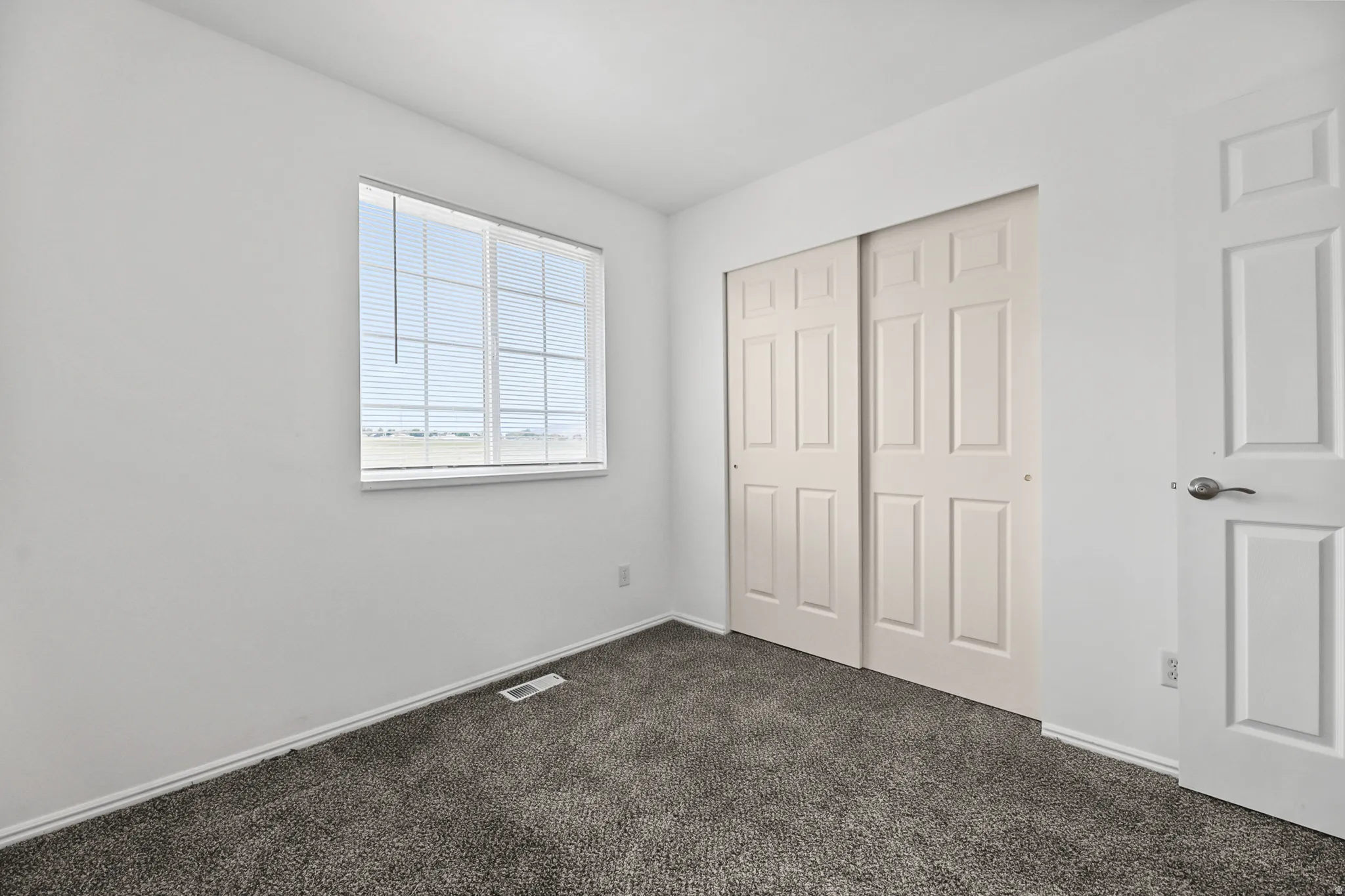 Room featuring a neutral color palette, grey textured carpeting, a white trim window with horizontal blinds, and a built-in sliding panel closet