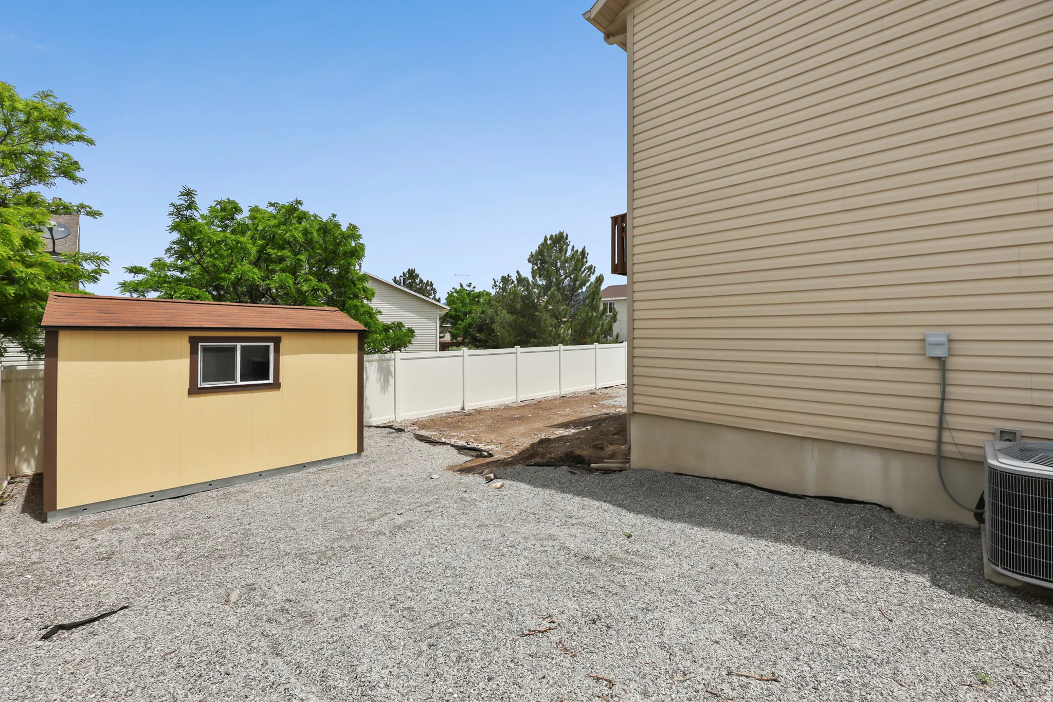 Gravel-covered side yard featuring a separate storage shed with a window, tan vinyl siding on the main structure, and a white privacy fence