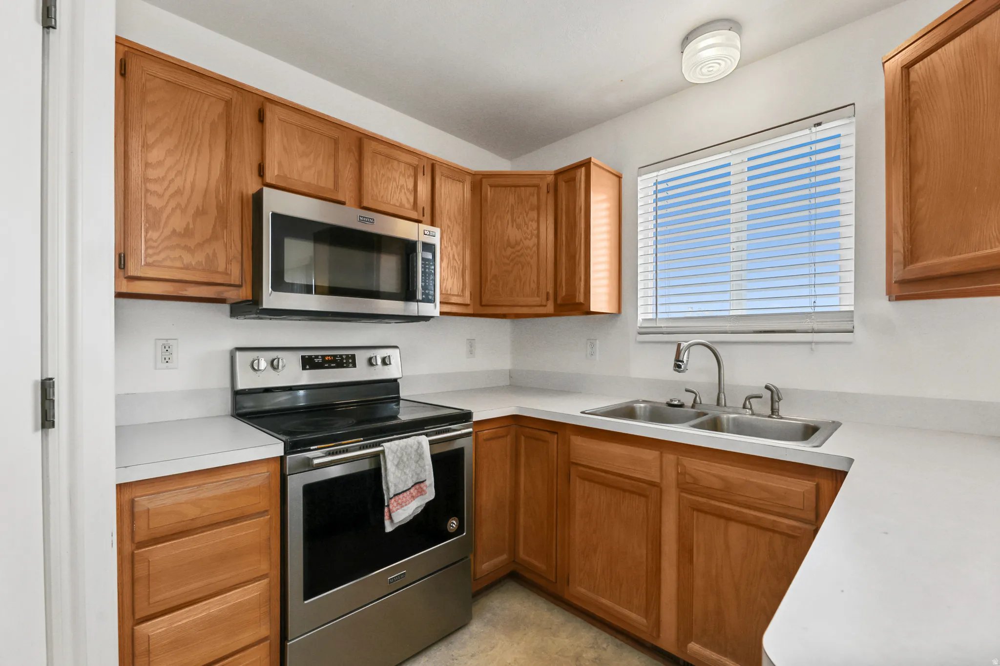 Kitchen featuring wood cabinetry, white countertops, a stainless steel range with overhead microwave, and a double basin sink beneath a window with blinds