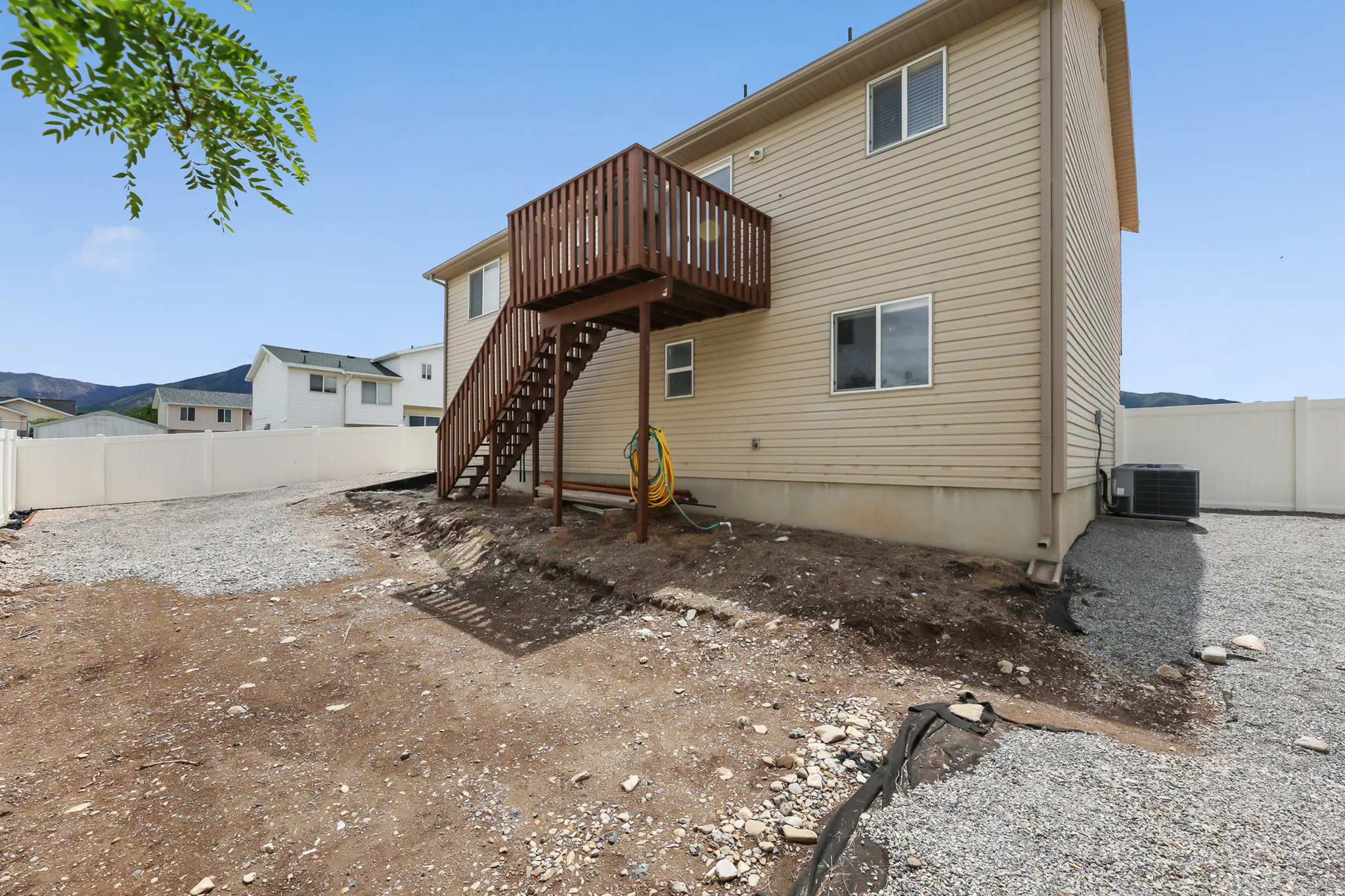 Rear exterior featuring a raised wood deck with staircase, beige siding, a white privacy fence, and a gravel yard