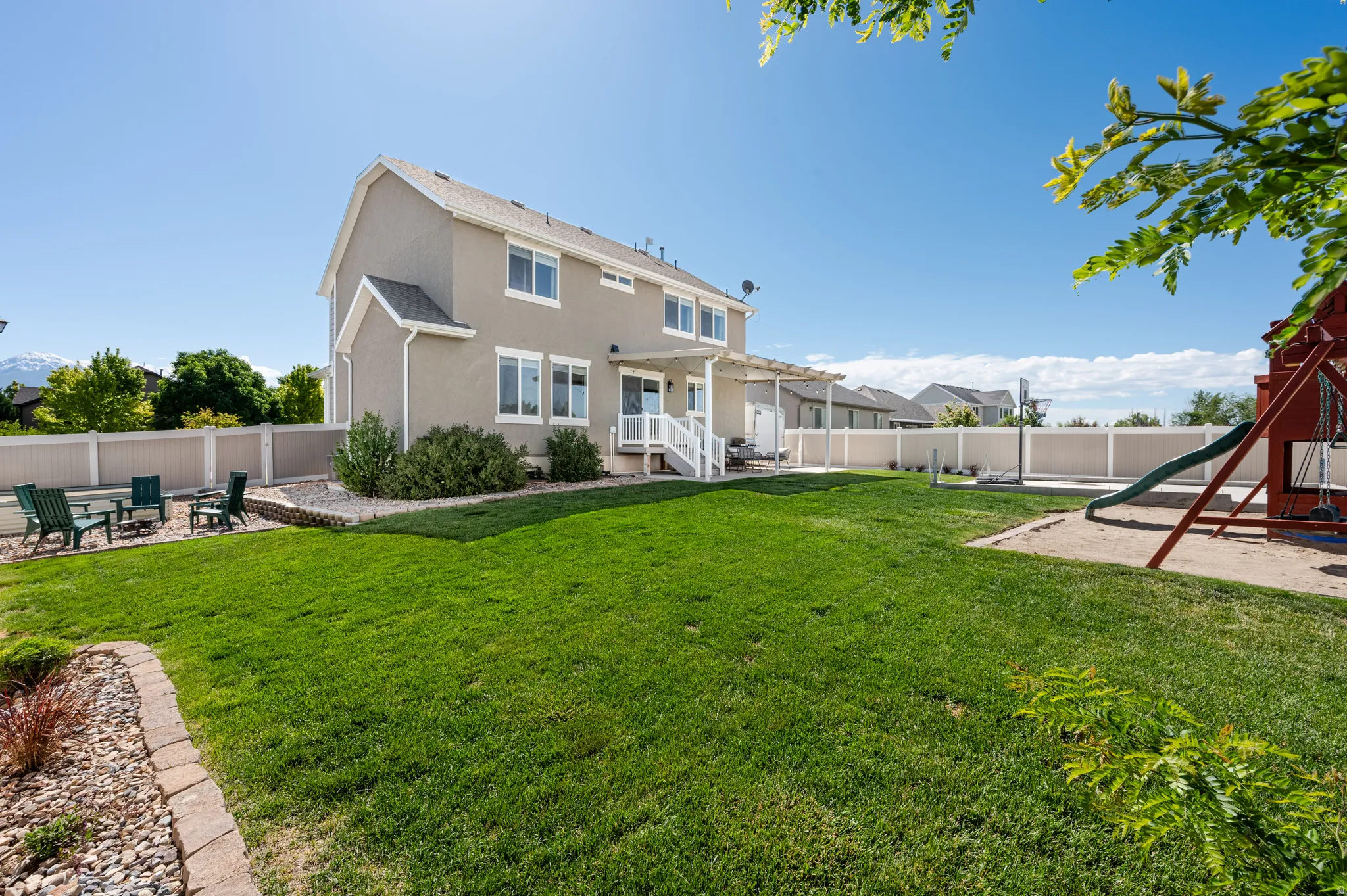 Expansive backyard featuring a manicured lawn, a gravel patio area with retaining wall, a white vinyl privacy fence, and a concrete patio with a pergola