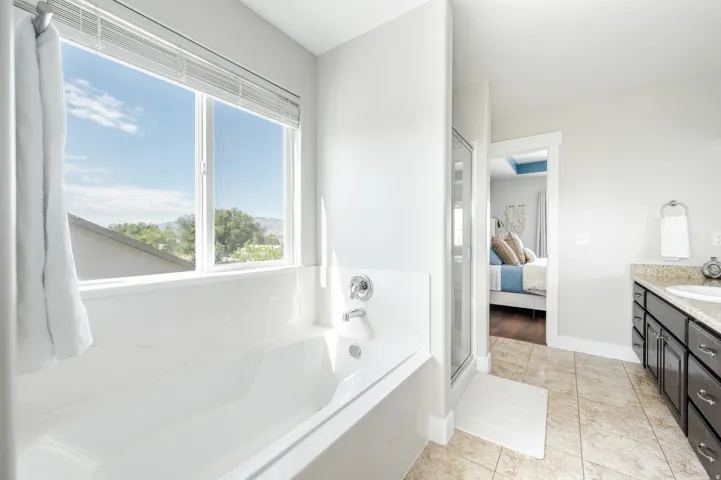 Soaking tub with a large window, tile flooring, glass-enclosed shower, and a vanity with a stone countertop