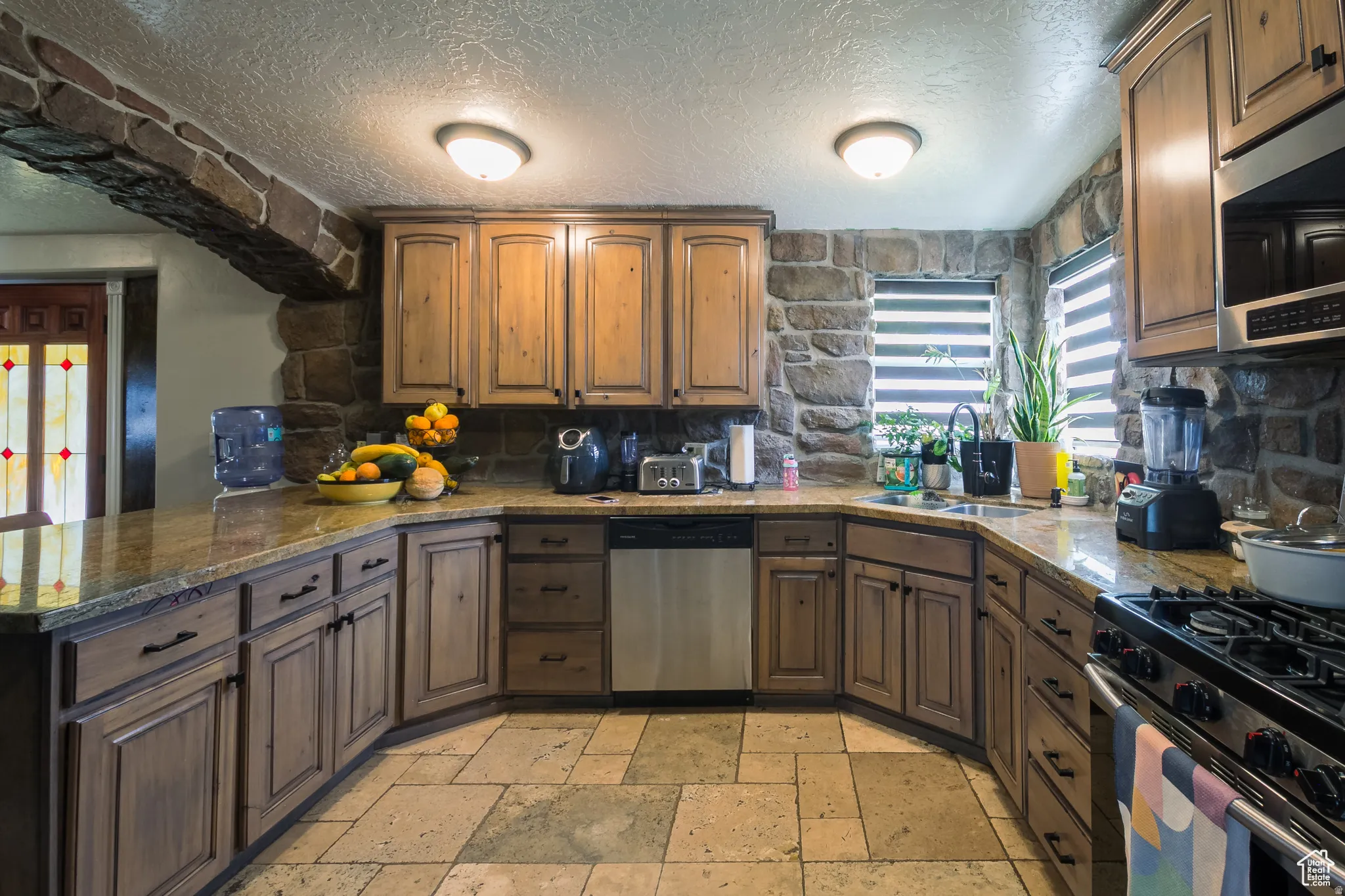 Kitchen featuring stone wall accents, wood-finish cabinetry, stone tile flooring, and integrated stainless steel appliances
