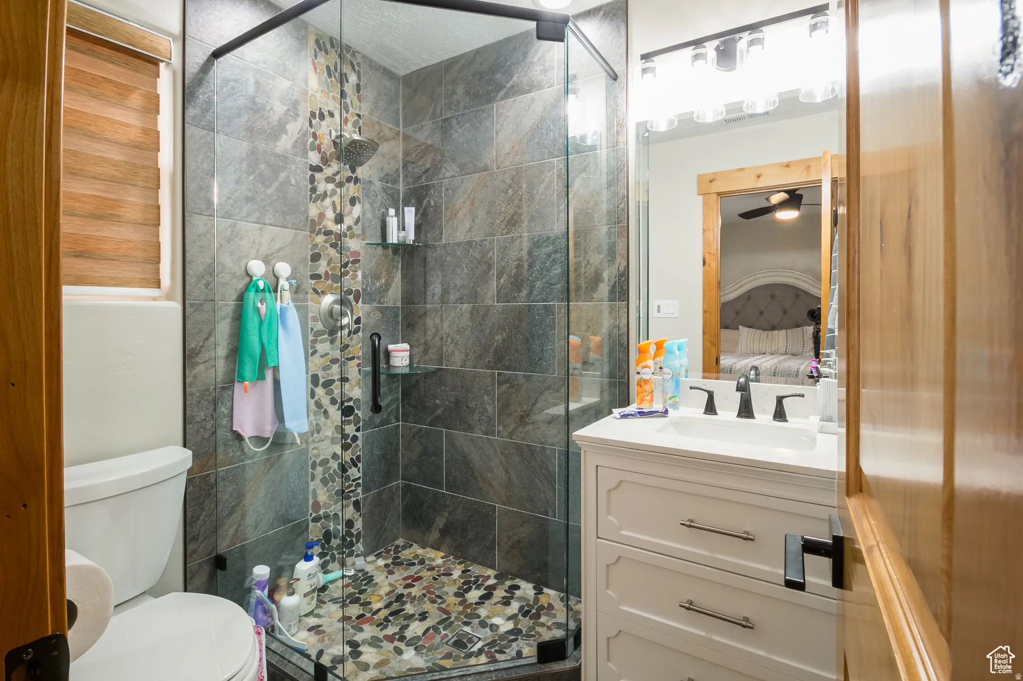 Walk-in shower with natural stone tile and river rock flooring, accompanied by a white vanity featuring a rectangular sink, dark fixtures, and a mirrored medicine cabinet