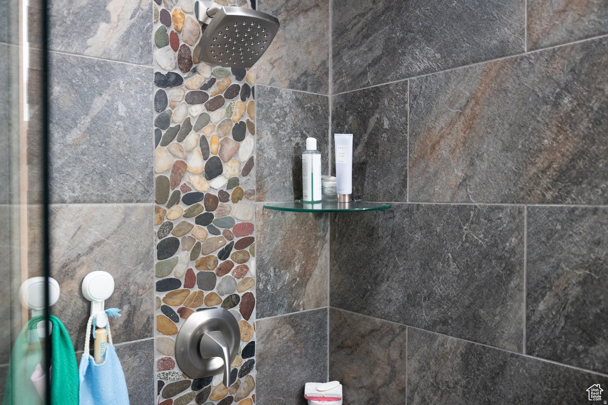 Shower featuring a pebbled vertical accent strip, large format stone tile, a brushed nickel showerhead, and a glass corner shelf