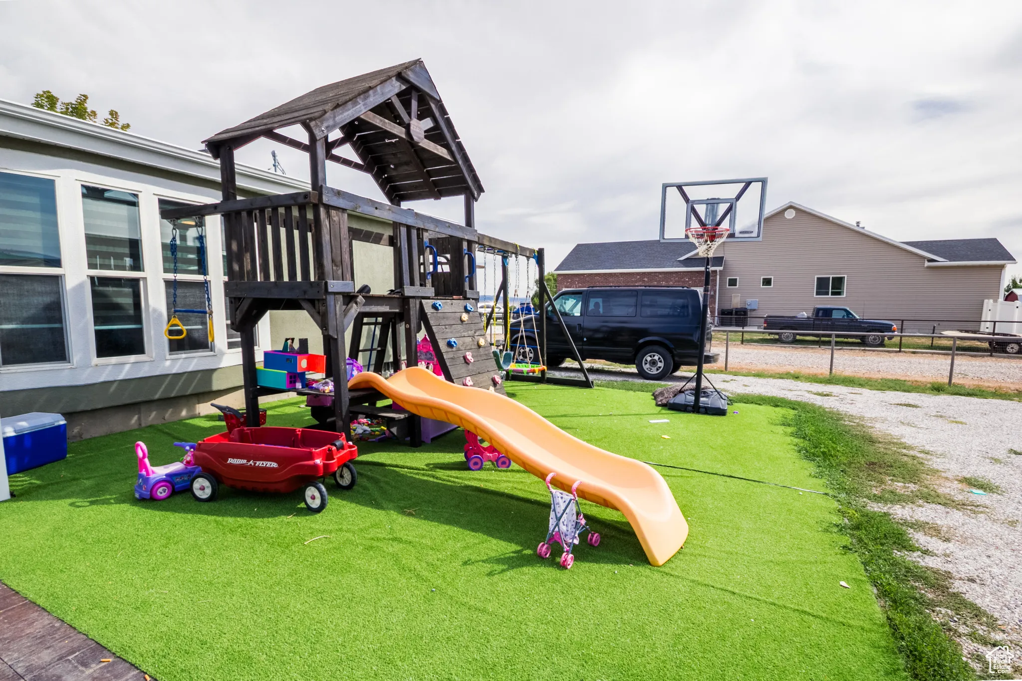Spacious outdoor area featuring a wooden playset with a slide, a basketball hoop, and artificial turf surfacing