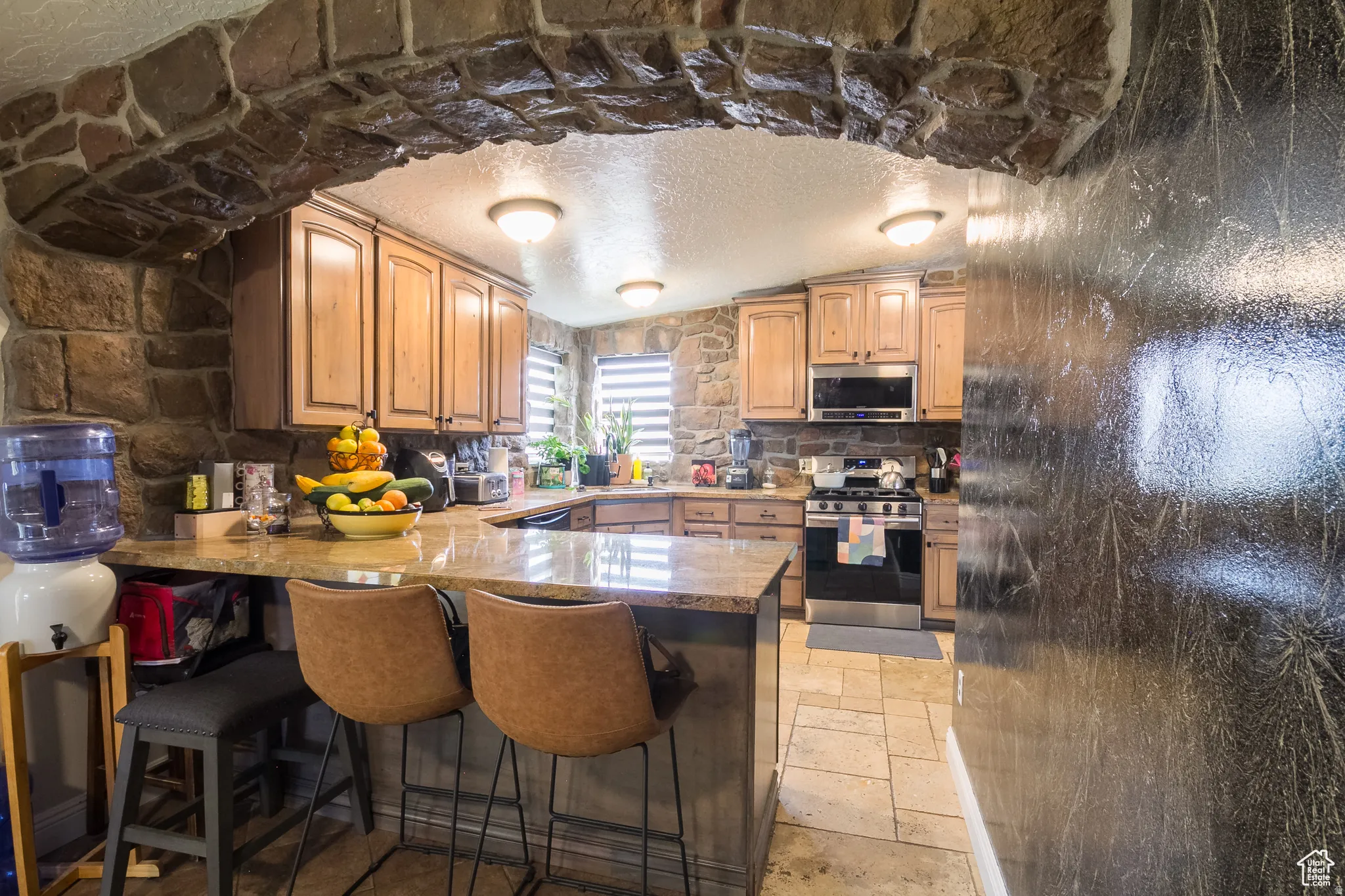Stone archway entry to a kitchen featuring wood cabinetry, stone-finish walls, a breakfast bar with stone-finish countertops, and stainless steel appliances