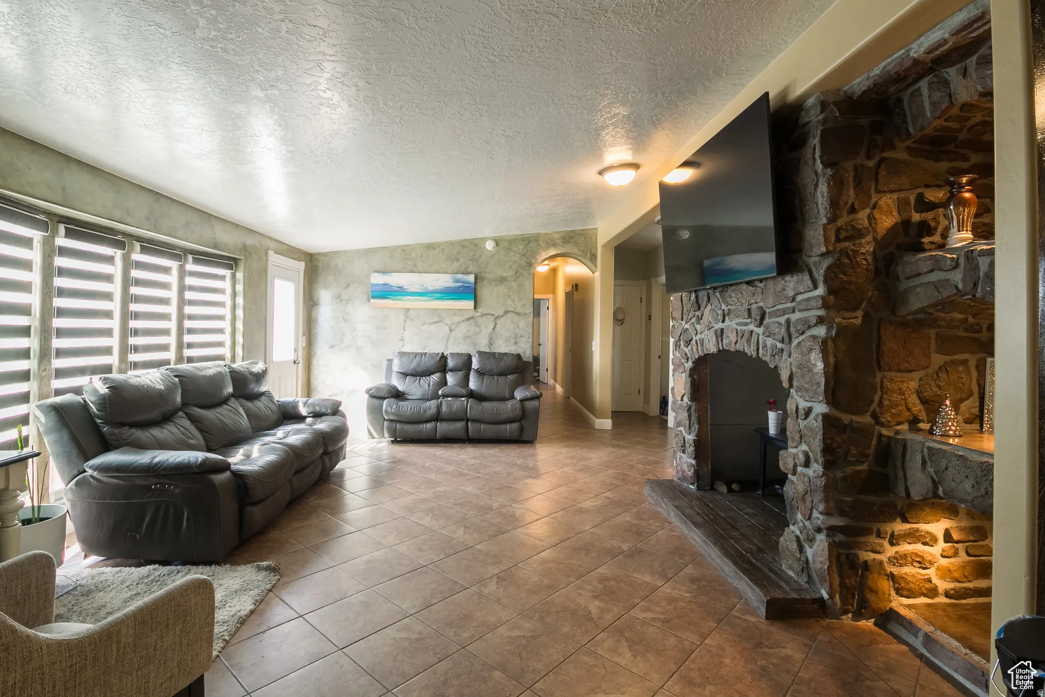 Spacious living area featuring a stone fireplace with integrated shelving, tile flooring, textured wall finishes, and multiple windows with horizontal blinds