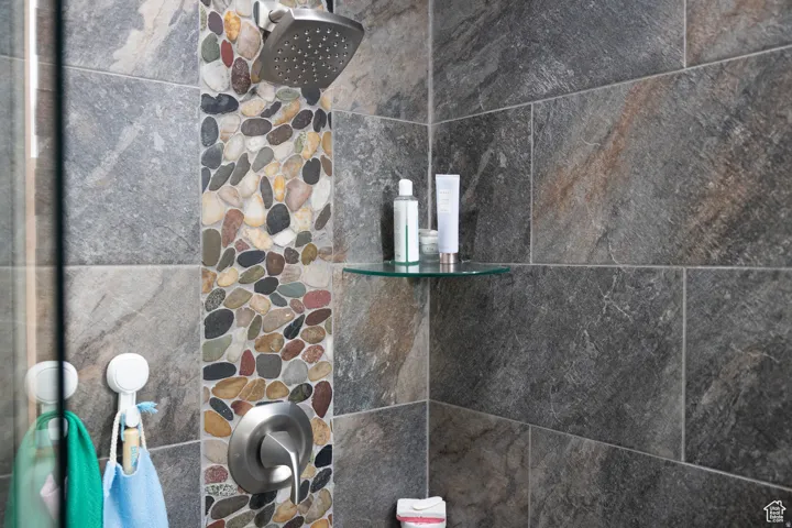 Shower featuring a pebbled vertical accent strip, large format stone tile, a brushed nickel showerhead, and a glass corner shelf