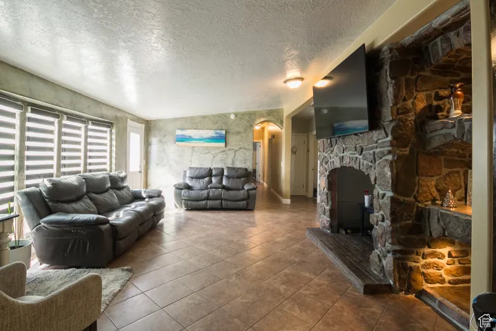 Spacious living area featuring a stone fireplace with integrated shelving, tile flooring, textured wall finishes, and multiple windows with horizontal blinds