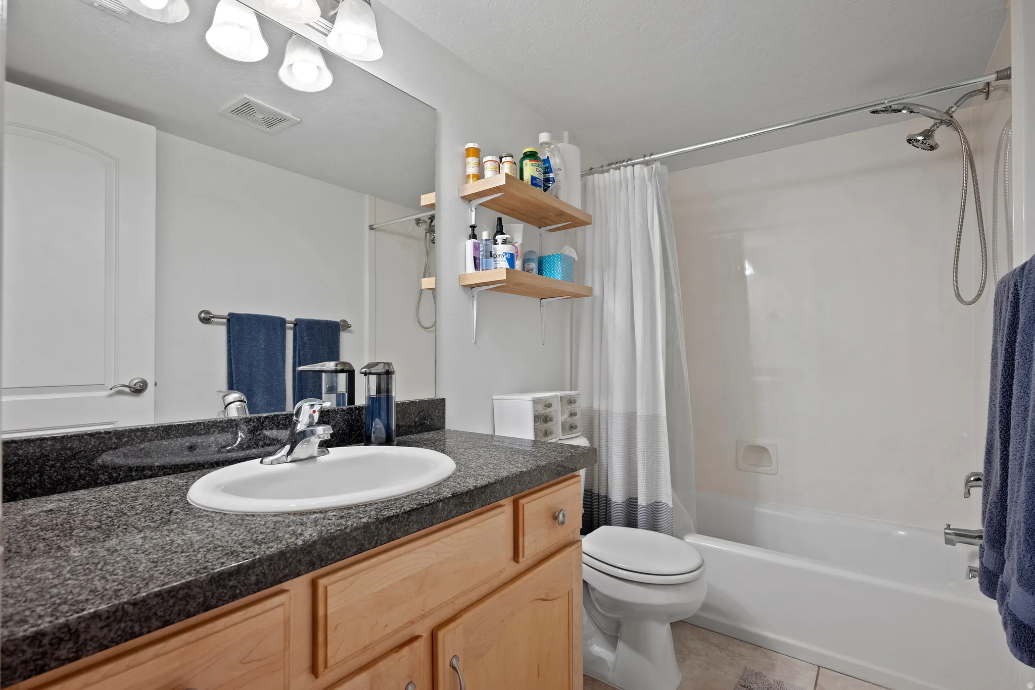 Bathroom featuring a granite-finish countertop, undermount sink, light wood-finish cabinetry, and a full-size tub with shower