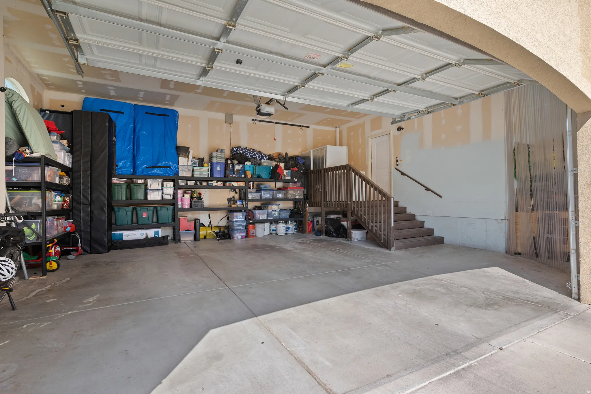 Spacious garage featuring concrete flooring, an overhead garage door, built-in shelving, and a wooden staircase with a handrail