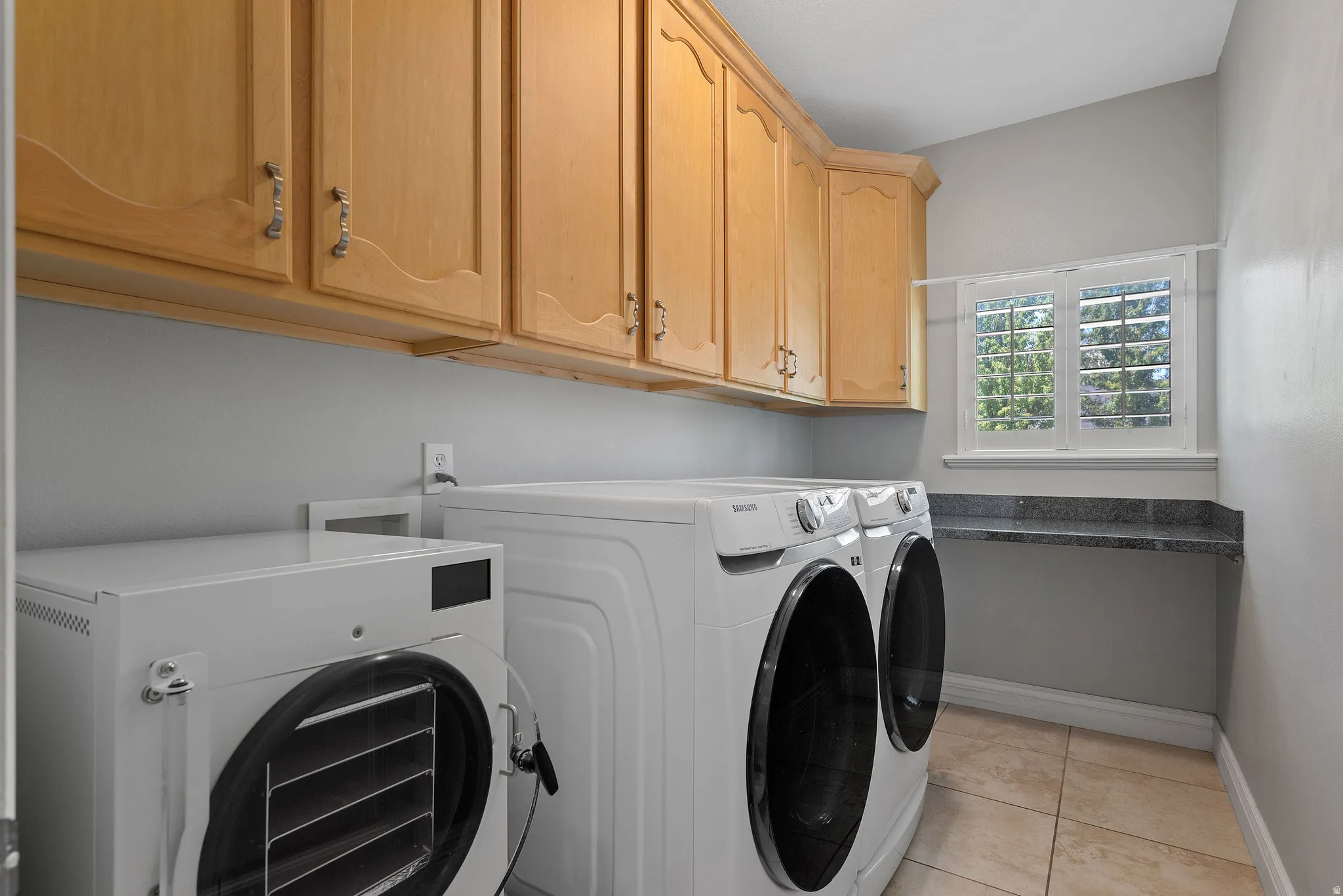 Laundry space featuring built-in cabinetry, tile flooring, a window with plantation shutters, and a granite-look countertop