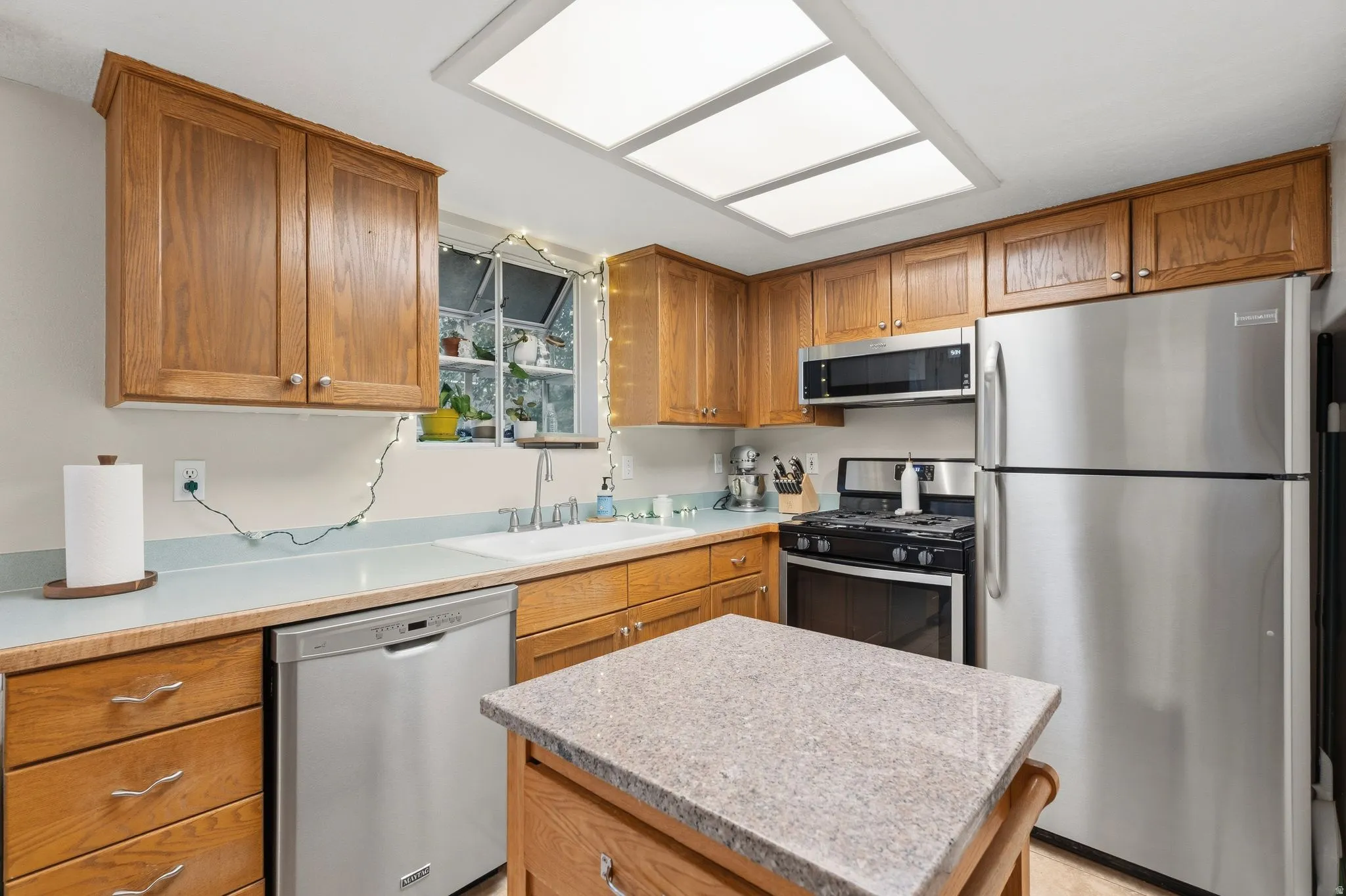 Kitchen featuring wood-finish cabinetry, stainless steel appliances, a center island with a stone-finish countertop, a single basin sink, and a multi-pane window