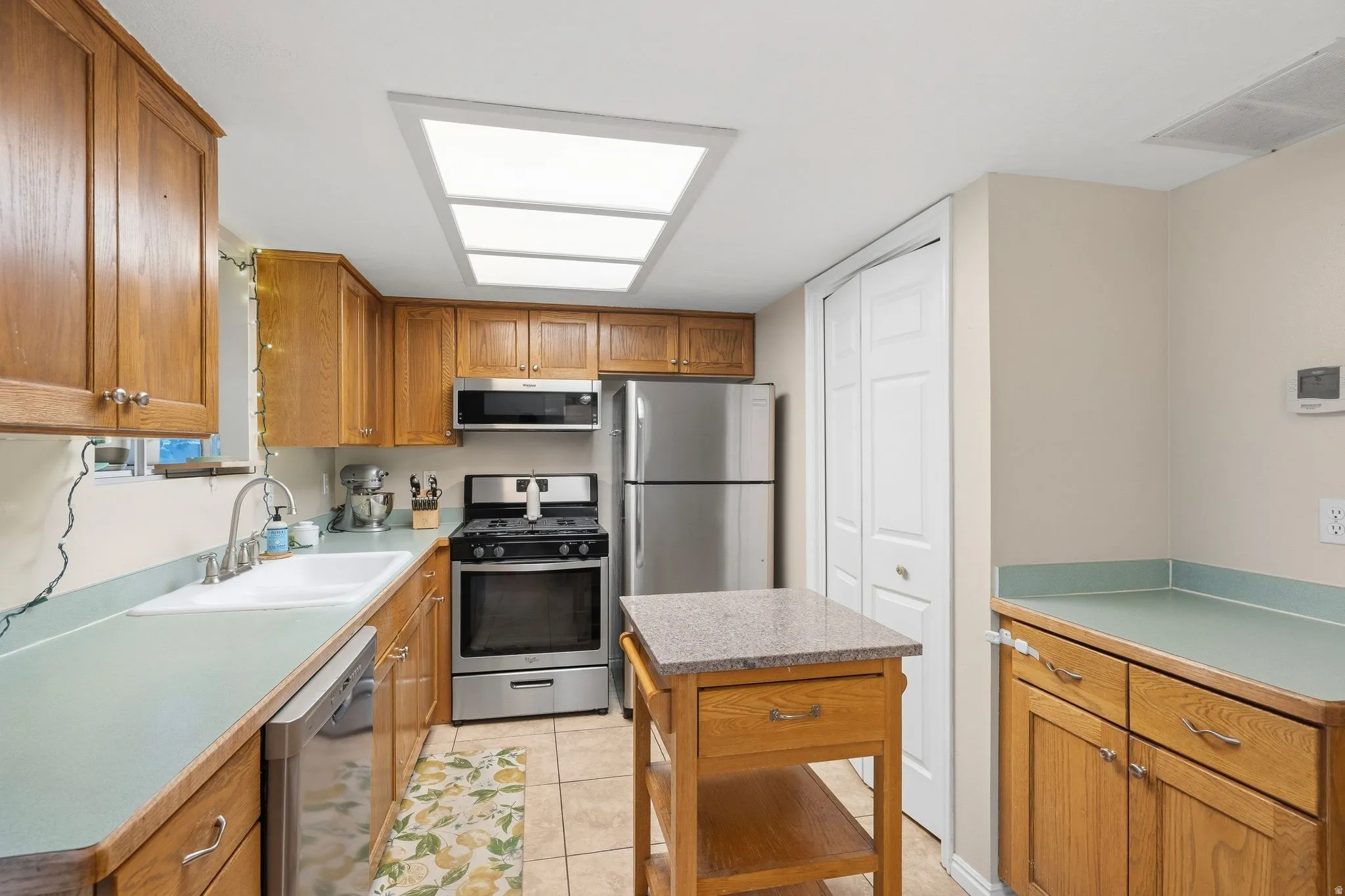 Kitchen featuring wood cabinetry, light-toned countertops, tile flooring, stainless steel appliances, and a central island with a granite-look top