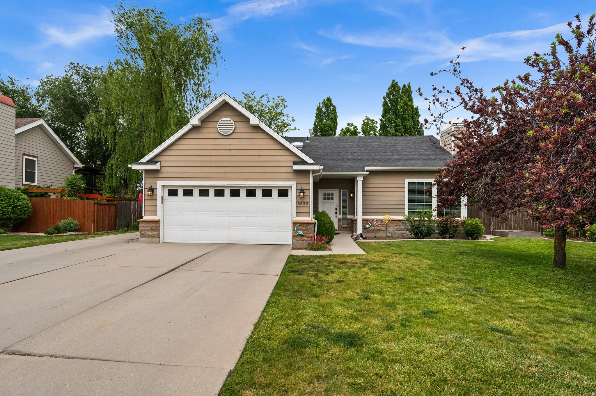 Ranch-style residence featuring a tan siding facade with stone veneer accents, a dark shingle roof, and a white garage door with rectangular window inserts