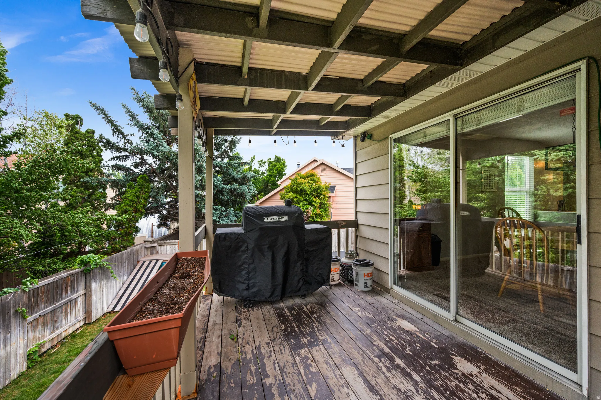 Expansive wood-finish deck with a corrugated metal patio cover, sliding glass door access, and string lighting