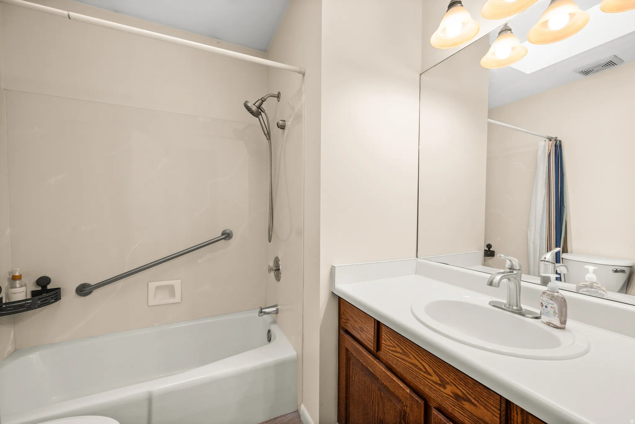 Bathroom featuring a white tub, shower with a handheld fixture, wall-mounted grab bar, and a single vanity with a white countertop and wood-finish cabinetry
