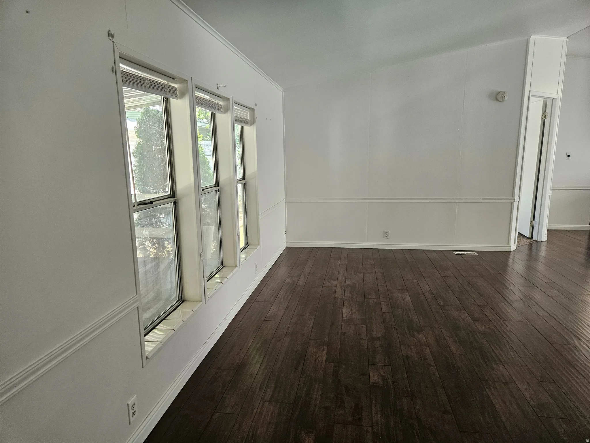 Open interior space featuring dark wood-finish flooring, white paneling, and a trio of windows providing natural illumination