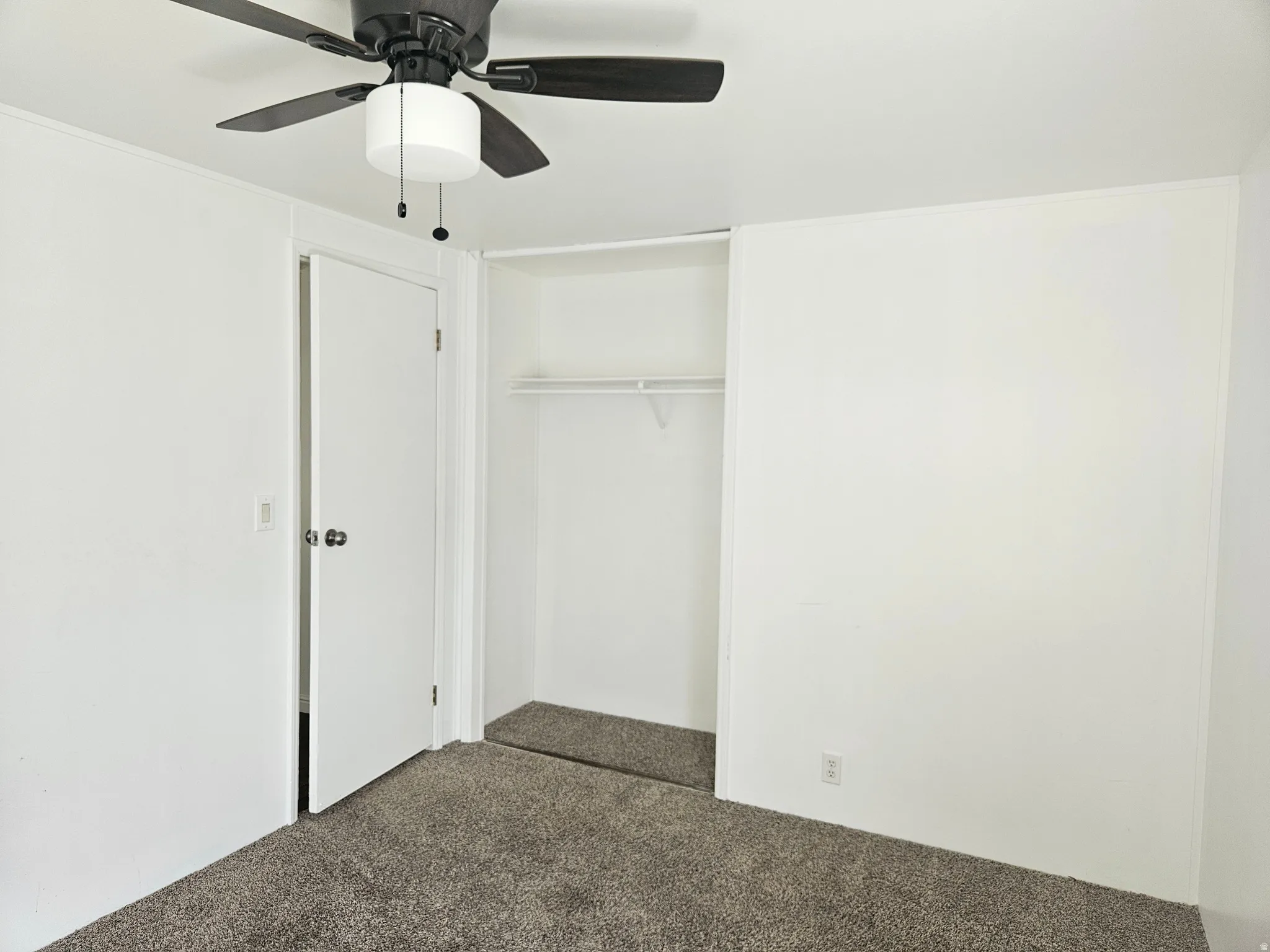 Interior room featuring neutral wall paint, dark grey carpet flooring, a ceiling fan with integrated lighting, and an open closet with shelving