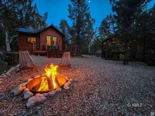 Gravel outdoor area featuring a stone-lined fire pit, two slatted wood chairs, and a wood-sided cabin with a deck