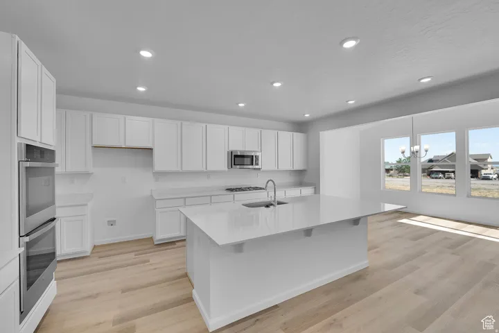 Open-concept kitchen featuring an oversized island with an undermount sink, light wood-finish flooring, white shaker cabinetry, stainless steel double wall ovens, and recessed lighting