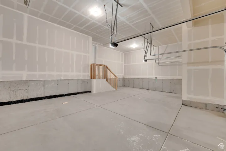 Spacious garage interior featuring a concrete floor, unfinished drywall, overhead lighting, garage door track system, and an interior staircase with wood railing