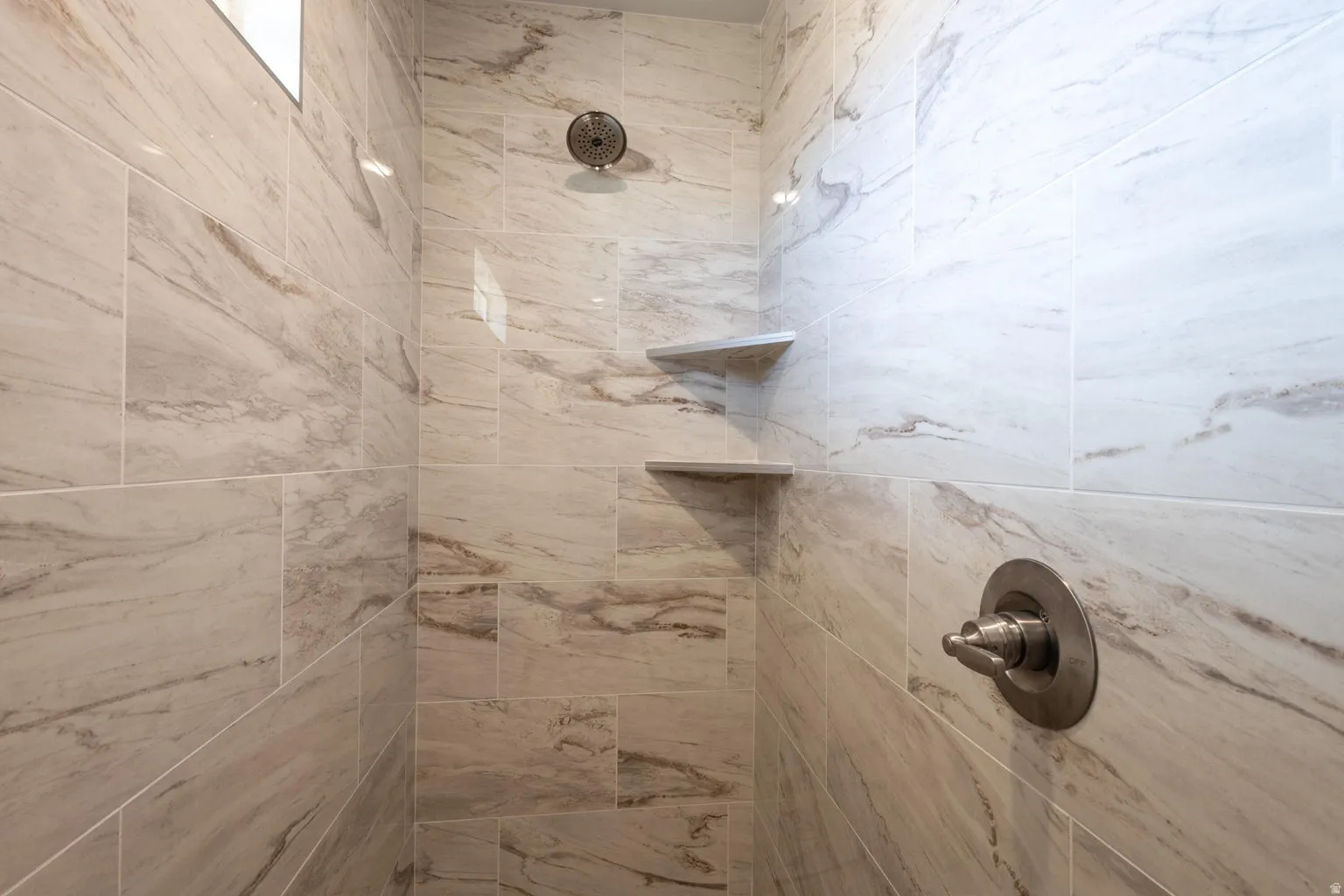 Shower interior featuring large-format tile with subtle veining, dual built-in shelving, a rainfall showerhead, and a single-handle control valve