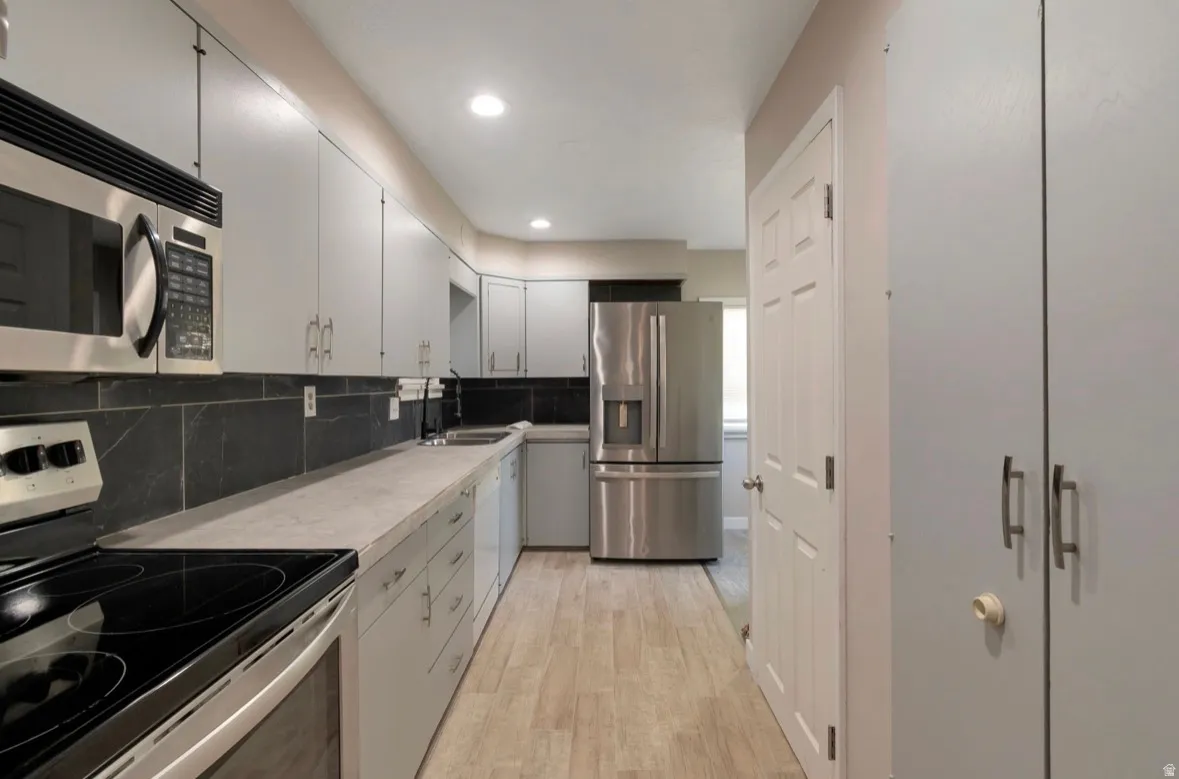 Galley kitchen featuring light gray cabinetry, stainless steel appliances, dark stone-look backsplash, light wood-finish flooring, and recessed lighting