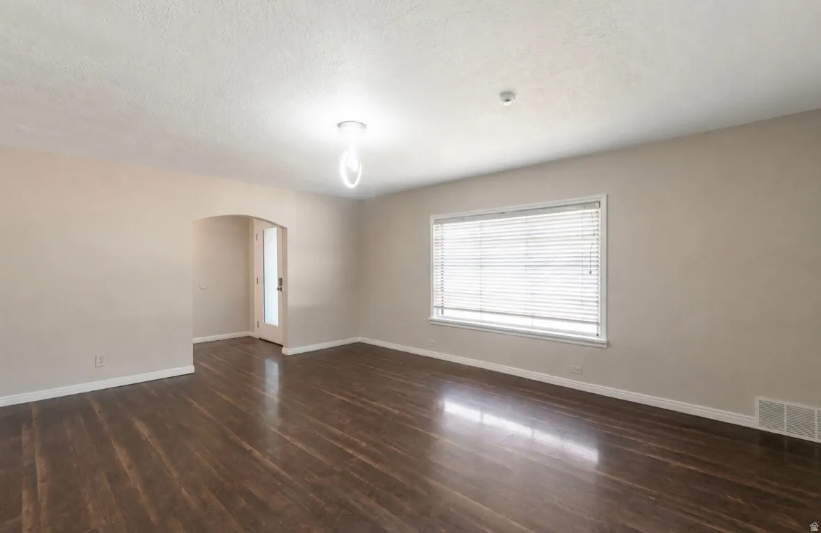 Spacious room featuring dark wood-finish flooring, neutral wall tones, a large window with horizontal blinds, and a contemporary ceiling fixture