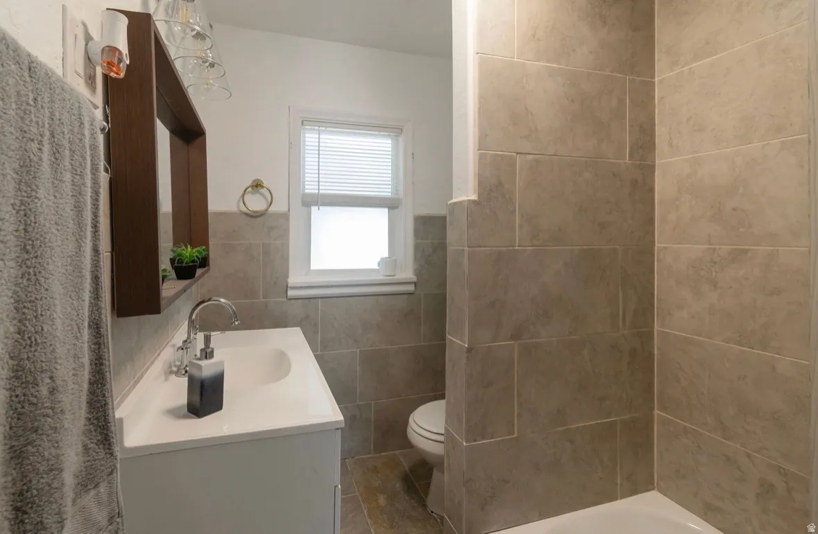 Bathroom featuring large format tile walls, a built-in tub with shower, a single vanity with a white countertop and integrated sink, and a white framed window with blinds