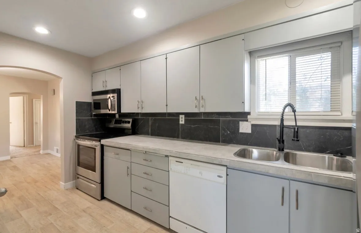 Kitchen featuring light gray cabinetry, stainless steel appliances, dark stone-look backsplash, dual basin sink with a matte black faucet, and wood-finish flooring