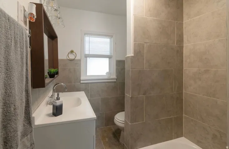 Bathroom featuring large format tile walls, a built-in tub with shower, a single vanity with a white countertop and integrated sink, and a white framed window with blinds