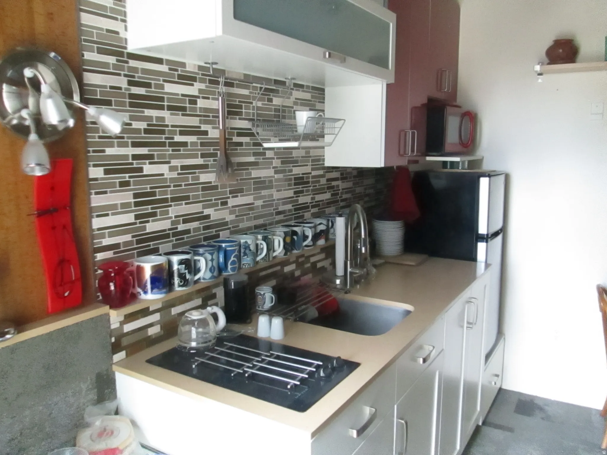 Compact kitchen with mosaic tile backsplash, integrated sink with gooseneck faucet, light-toned countertop, and white cabinetry with brushed nickel hardware