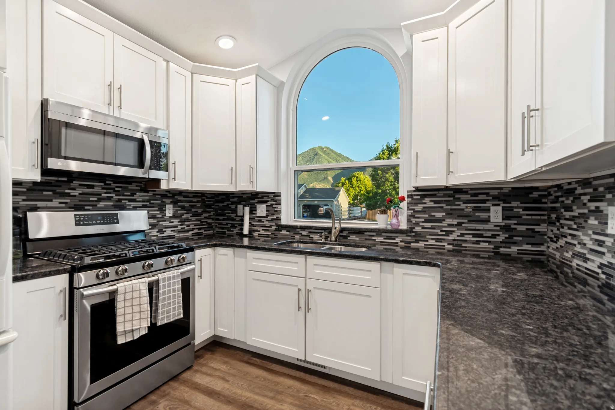 Kitchen featuring a large arched window, white cabinetry with brushed nickel hardware, stainless steel appliances, dark stone countertops, and a multi-tone tile backsplash