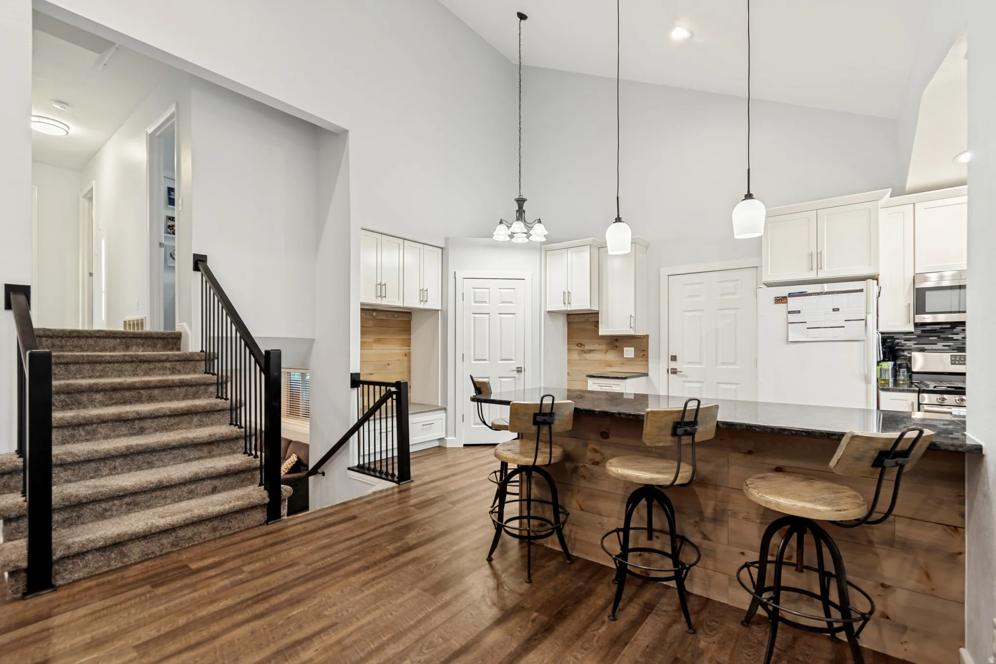 Open-concept living space with vaulted ceilings, wood-finish flooring, a carpeted staircase with black iron railings, and a kitchen featuring a wood-paneled island with a stone countertop, pendant lighting, and white cabinetry