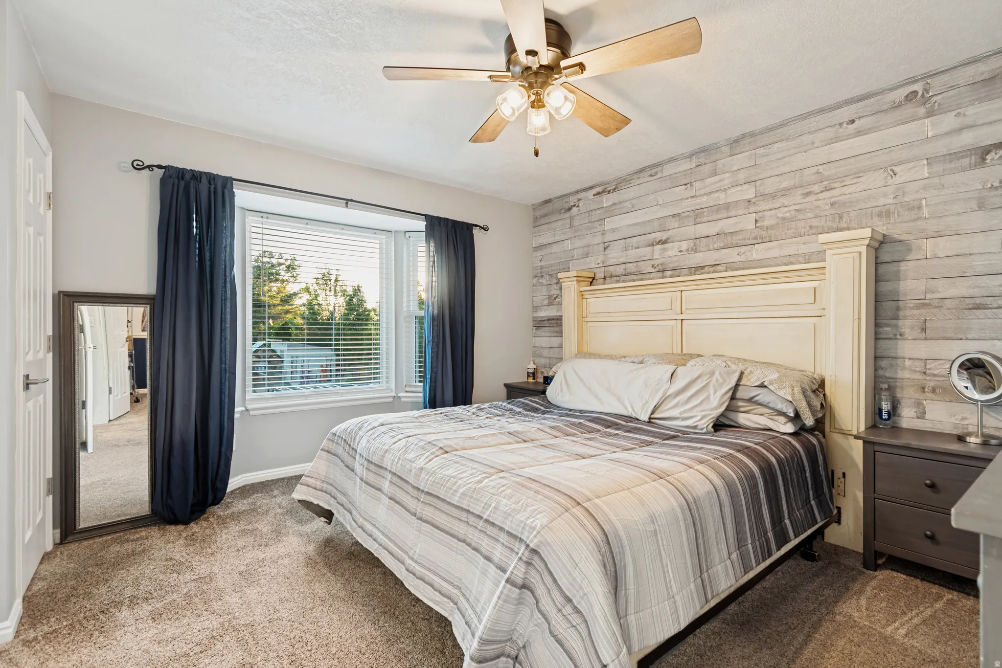 Carpeted room featuring a bay window with blinds, a wood-finish accent wall, and a ceiling fan with integrated lighting