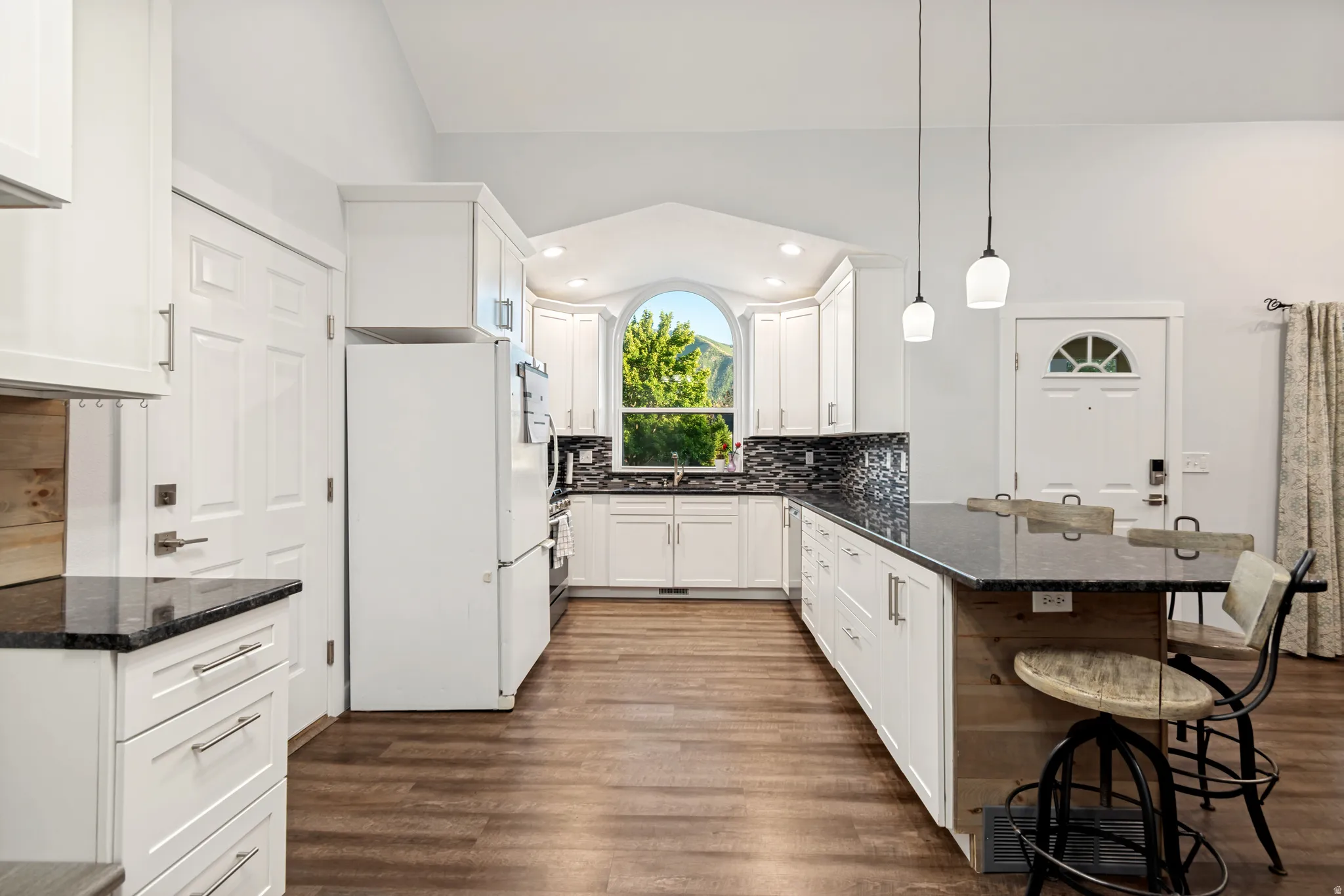 Bright kitchen featuring white cabinetry, dark countertops, a mosaic tile backsplash, wood-finish flooring, and a large arched window