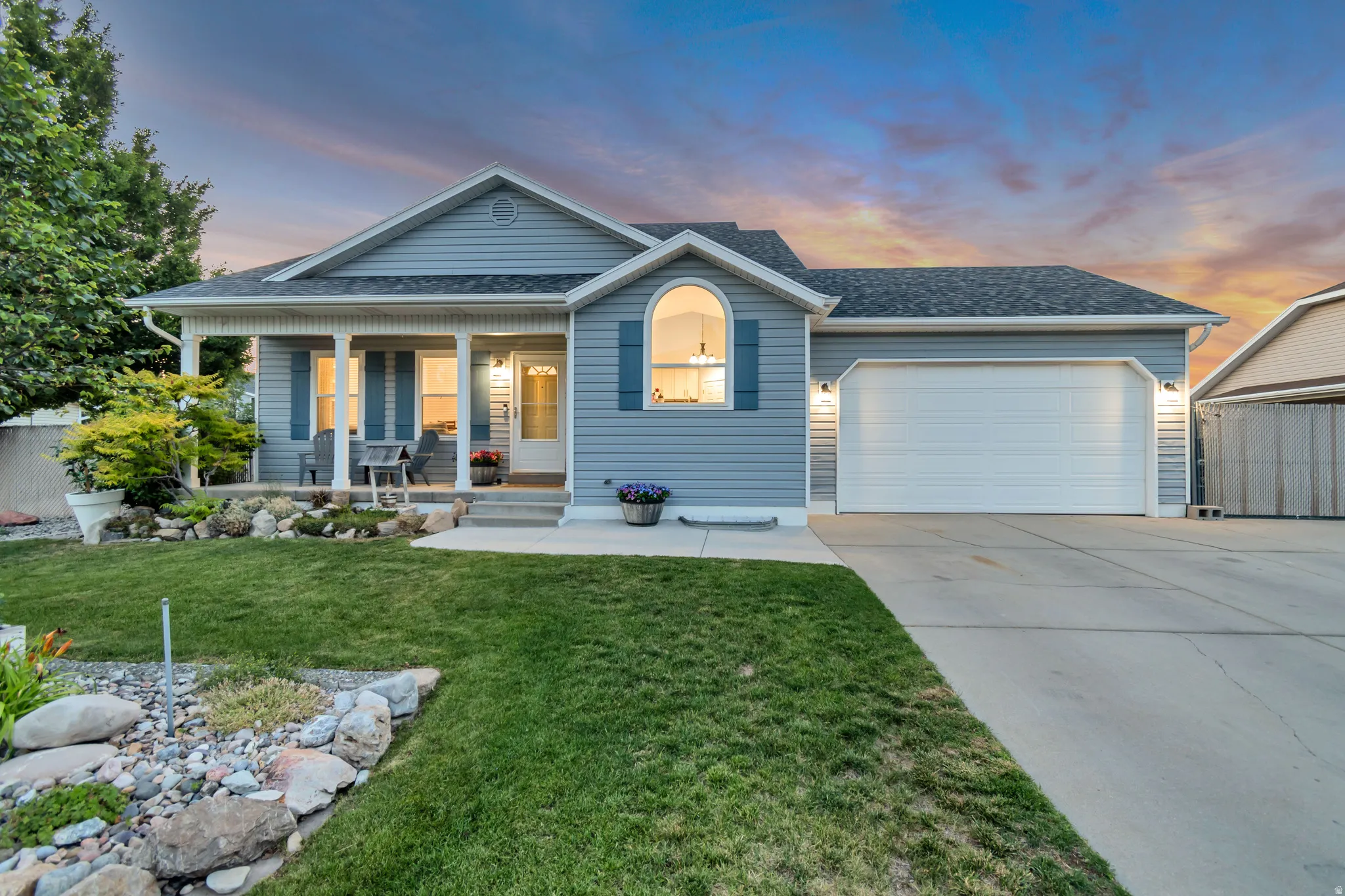Exterior featuring light blue siding with white trim, a covered front porch with white columns, a two-car garage with a white door, a manicured lawn, and a decorative rock garden